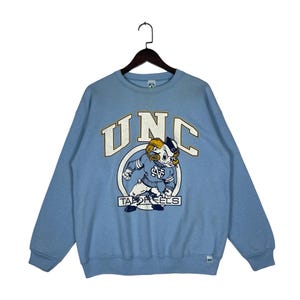 May include: A light blue crewneck sweatshirt featuring the text UNC in large white letters with a gold outline. Below the text is a graphic of a ram mascot wearing a blue sweater with the letters NC, positioned above the text TAR HEELS.