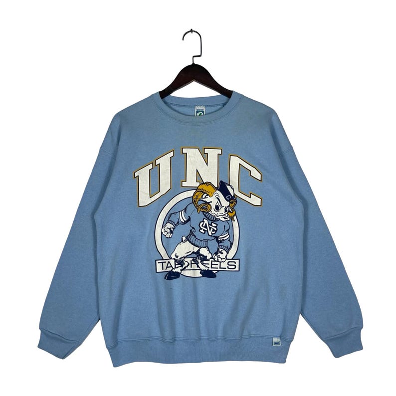 May include: A light blue crewneck sweatshirt featuring the text UNC in large white letters with a gold outline. Below the text is a graphic of a ram mascot wearing a blue sweater with the letters NC, positioned above the text TAR HEELS.