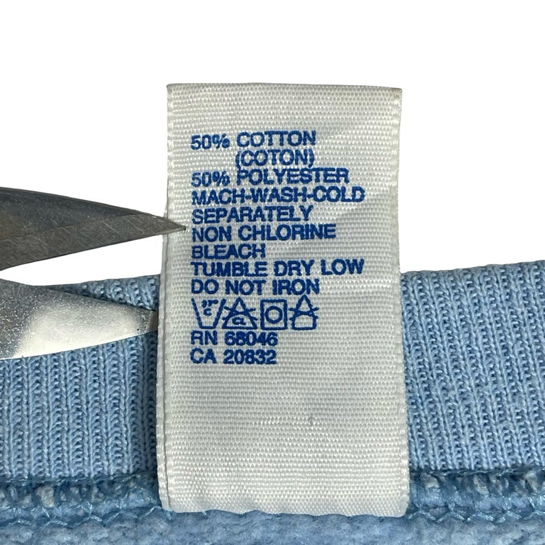 May include: A white fabric care label attached to a light blue garment. The label features blue text stating 50% COTTON, 50% POLYESTER, MACH-WASH-COLD, SEPARATELY, NON CHLORINE BLEACH, TUMBLE DRY LOW, DO NOT IRON, RN 68046, CA 20832, and laundry symbols.