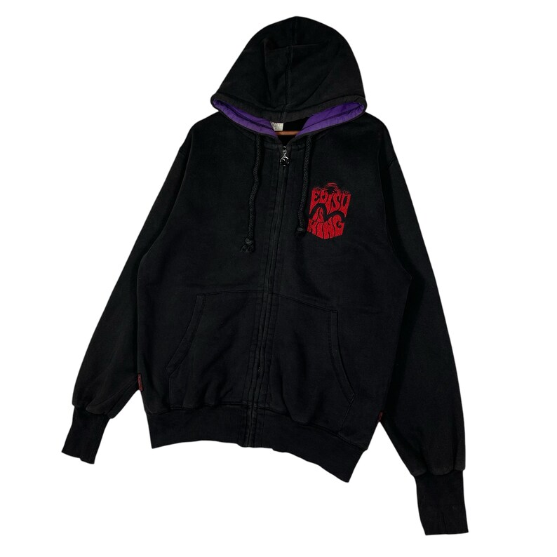 May include: Black zip-up hoodie with a purple-lined hood and cuffs. The hoodie features a red graphic on the chest. The hoodie has a drawstring and two front pockets.