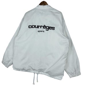 May include: White Courr&egrave;ges sport jacket with black lettering on the back. The jacket has a drawstring at the bottom and a collar. The jacket is hanging on a wooden hanger.