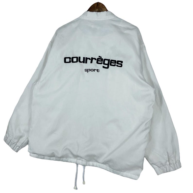 May include: White Courr&egrave;ges sport jacket with black lettering on the back. The jacket has a drawstring at the bottom and a collar. The jacket is hanging on a wooden hanger.
