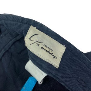 May include: Close-up of navy blue fabric with a white label. The label has the text "G/S Cottoning Workshop" in black. A smaller white label with the text "JAPAN" is also visible. The fabric appears to be part of a garment.