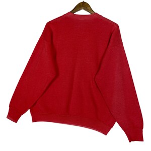 May include: A red sweatshirt hangs from a wooden hanger against a white background. The sweatshirt has a crew neck and long sleeves. The fabric appears to be a soft, knit material. The overall style is casual and comfortable.