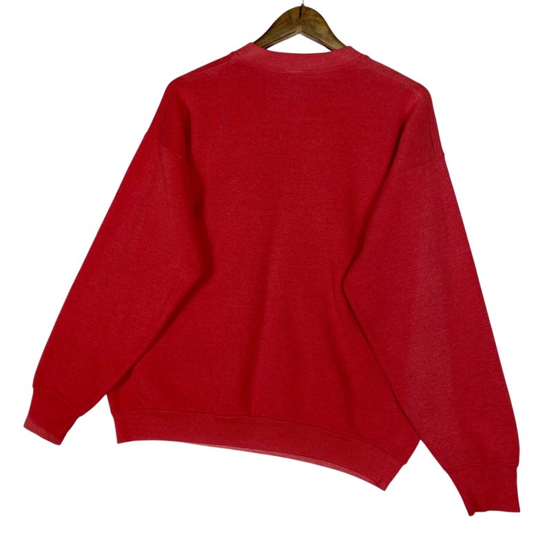 May include: A red sweatshirt hangs from a wooden hanger against a white background. The sweatshirt has a crew neck and long sleeves. The fabric appears to be a soft, knit material. The overall style is casual and comfortable.