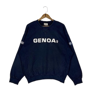 May include: Navy blue crew neck sweatshirt with the word "GENOA" in white block letters across the chest. The sweatshirt has a small patch on the left sleeve and a crest on the right sleeve. The garment is hanging on a wooden hanger.