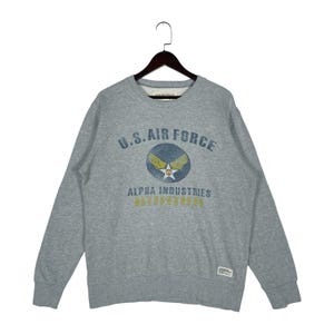 May include: A grey crew neck sweatshirt displaying the text "U.S. AIR FORCE" in blue, along with a circular emblem featuring wings and a star. The sweatshirt also has the text "ALPHA INDUSTRIES" in yellow. It is suspended from a wooden hanger.