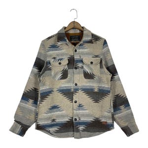 May include: A button-down shirt with a Southwestern-inspired geometric pattern in shades of beige, gray, blue, and brown. The shirt has two chest pockets and the brand name "JACKS" is visible on the label.