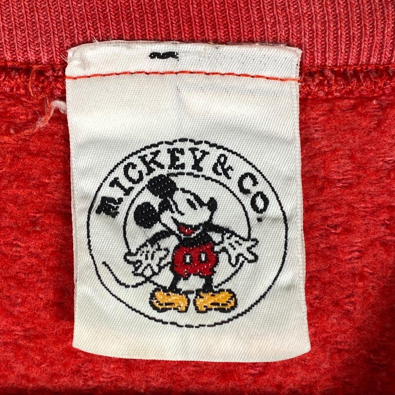 May include: Close-up of a red fabric with a white label featuring a Mickey Mouse graphic. The label reads "MICKEY & CO" in a circular design. The Mickey Mouse illustration is in black, red, yellow, and white.