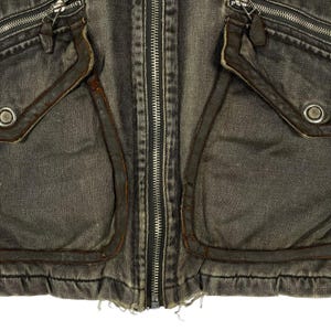 May include: Close-up of a distressed denim jacket in grey and brown tones. The jacket has a full-length silver zip, two front pockets with leather trim, and silver eyelets. The fabric has a worn, vintage appearance.