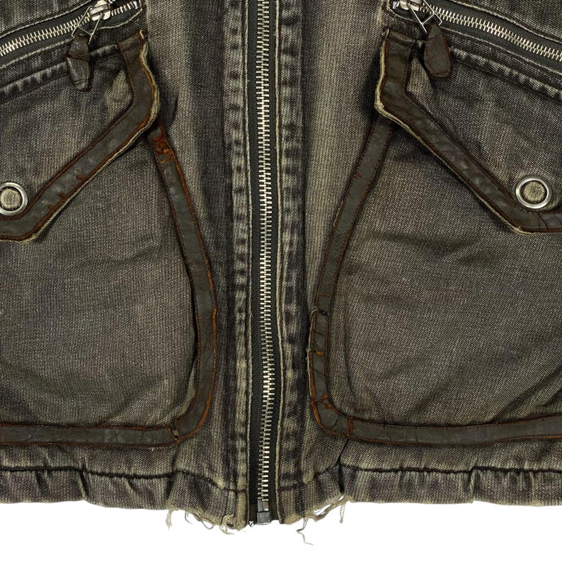 May include: Close-up of a distressed denim jacket in grey and brown tones. The jacket has a full-length silver zip, two front pockets with leather trim, and silver eyelets. The fabric has a worn, vintage appearance.