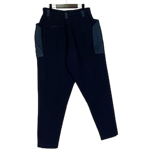 May include: Dark navy blue pants with two large, square pockets on the sides. The pants are hanging on a wooden hanger. The pockets are a lighter shade of blue. The pants have a high waist and a straight leg.
