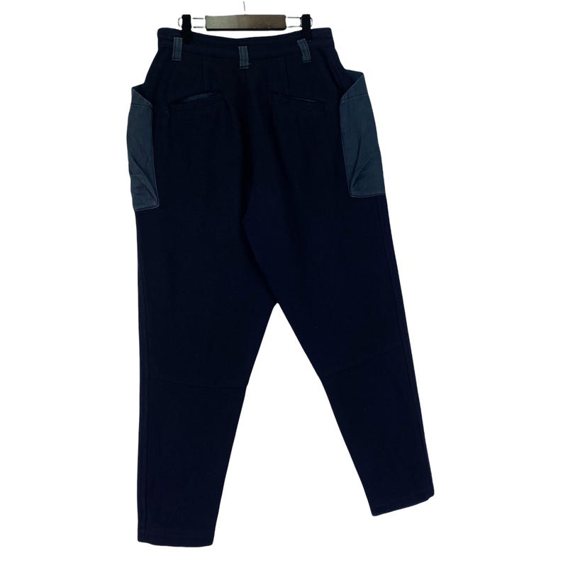 May include: Dark navy blue pants with two large, square pockets on the sides. The pants are hanging on a wooden hanger. The pockets are a lighter shade of blue. The pants have a high waist and a straight leg.