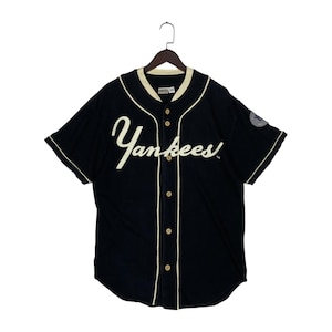 Könnte beinhalten: Ein schwarzes kurzärmeliges Baseballtrikot mit dem Schriftzug Yankees in weißer Schreibschrift auf der Brust. Das geknöpfte Hemd hat cremefarbene Paspeln am Kragen und an der Knopfleiste sowie ein rundes Team-Logo-Emblem am linken Ärmel.