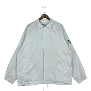 May include: White coach jacket with a collared neckline, snap-button closure, and long sleeves. Features two front pockets and a drawstring hem. The left sleeve has a black logo. The jacket is hanging on a wooden hanger.