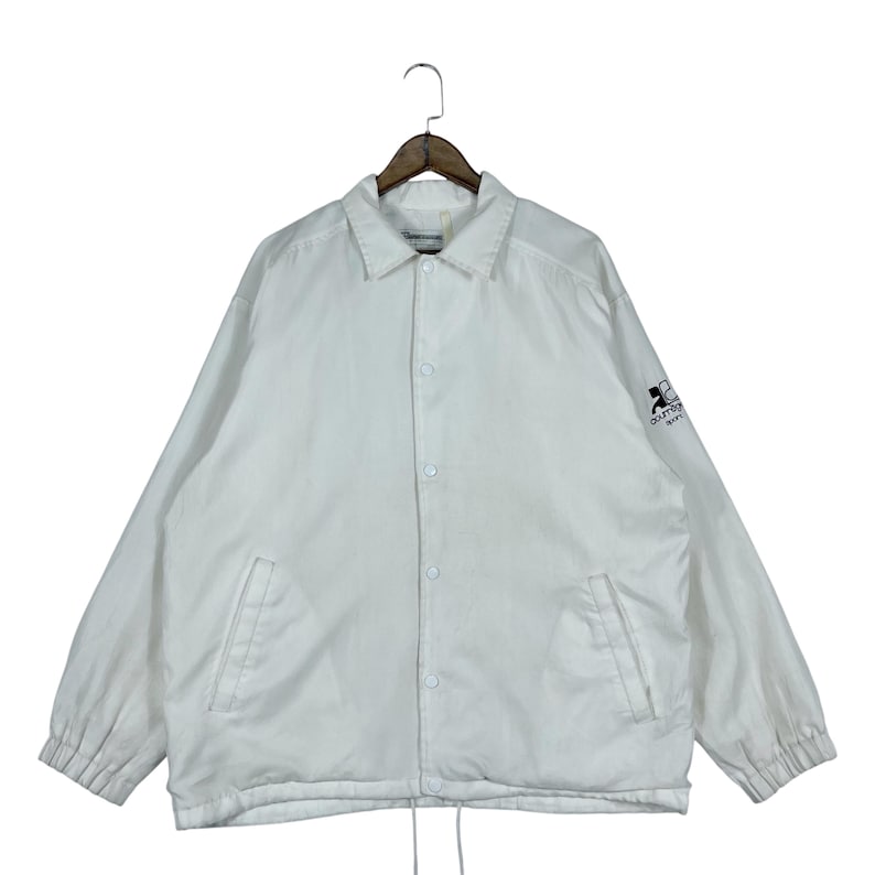May include: White coach jacket with a collared neckline, snap-button closure, and long sleeves. Features two front pockets and a drawstring hem. The left sleeve has a black logo. The jacket is hanging on a wooden hanger.