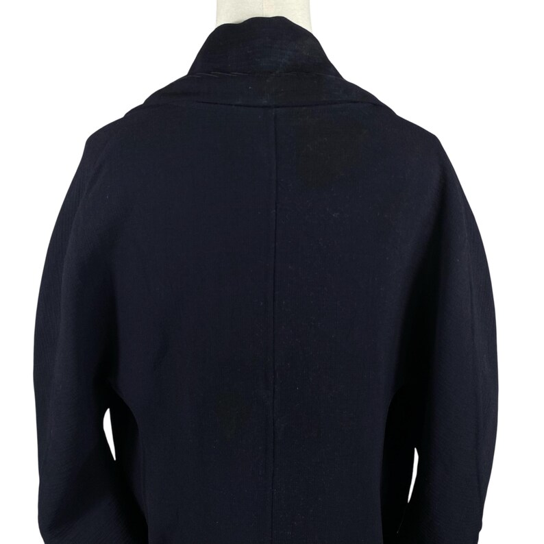 May include: A navy blue coat with a wide collar and a tailored fit. The back of the coat is visible, showcasing the clean lines and structured design. The coat is displayed on a mannequin, highlighting its shape and style.