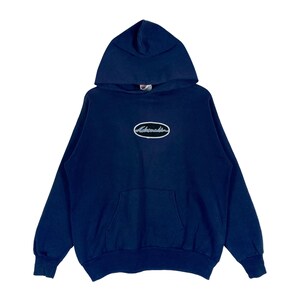 Puede incluir: Una sudadera con capucha de color azul marino oscuro que presenta un bolsillo tipo canguro frontal y un parche ovalado central con el texto Adrenalin bordado en blanco. La prenda tiene mangas largas con puños y dobladillo inferior acanalados.