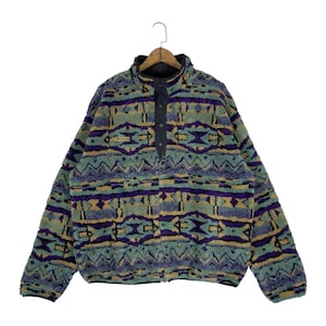 May include: A patterned fleece pullover in shades of green, purple, and tan. The long-sleeved top features a high collar and a button-up placket. The geometric design covers the entire garment, creating a cozy and stylish look.
