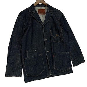 Puede incluir: Chaqueta vaquera azul oscuro con dise&ntilde;o de blazer. Presenta cuello con solapas, cierre de botones y m&uacute;ltiples bolsillos. La chaqueta est&aacute; colgada en una percha de madera. Una etiqueta roja es visible en el bolsillo.
