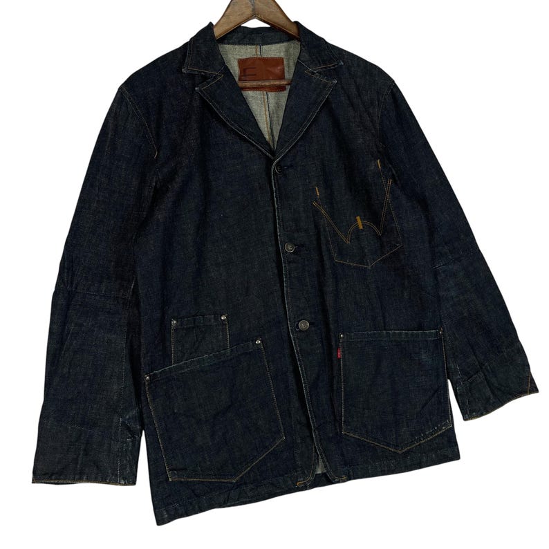 Puede incluir: Chaqueta vaquera azul oscuro con dise&ntilde;o de blazer. Presenta cuello con solapas, cierre de botones y m&uacute;ltiples bolsillos. La chaqueta est&aacute; colgada en una percha de madera. Una etiqueta roja es visible en el bolsillo.