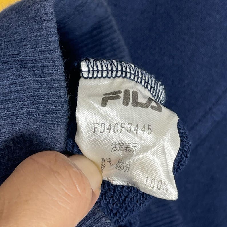 May include: A navy blue garment featuring a white interior fabric care label. The label displays the FILA brand name in black text, the alphanumeric code FD4CF3445, and Japanese characters. The fabric has a ribbed texture along the top edge.