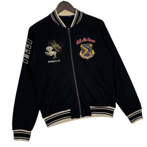 May include: Black bomber jacket with white and cream striped trim. Features embroidered dragon and U.S. Air Force patches. The jacket has a zip closure and the text "AVIREX LTD" on the front.