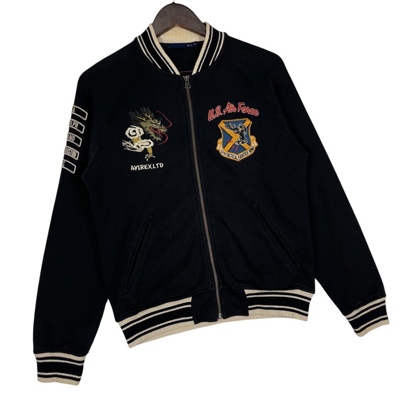 May include: Black bomber jacket with white and cream striped trim. Features embroidered dragon and U.S. Air Force patches. The jacket has a zip closure and the text "AVIREX LTD" on the front.