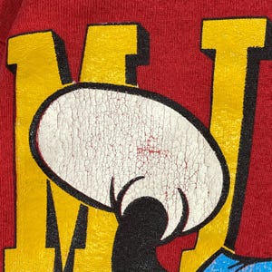 May include: Close-up of a red t-shirt featuring a distressed graphic of a cartoon character's face. The character's face is white with black details, set against yellow and red block letters. The shirt's fabric has a textured appearance.
