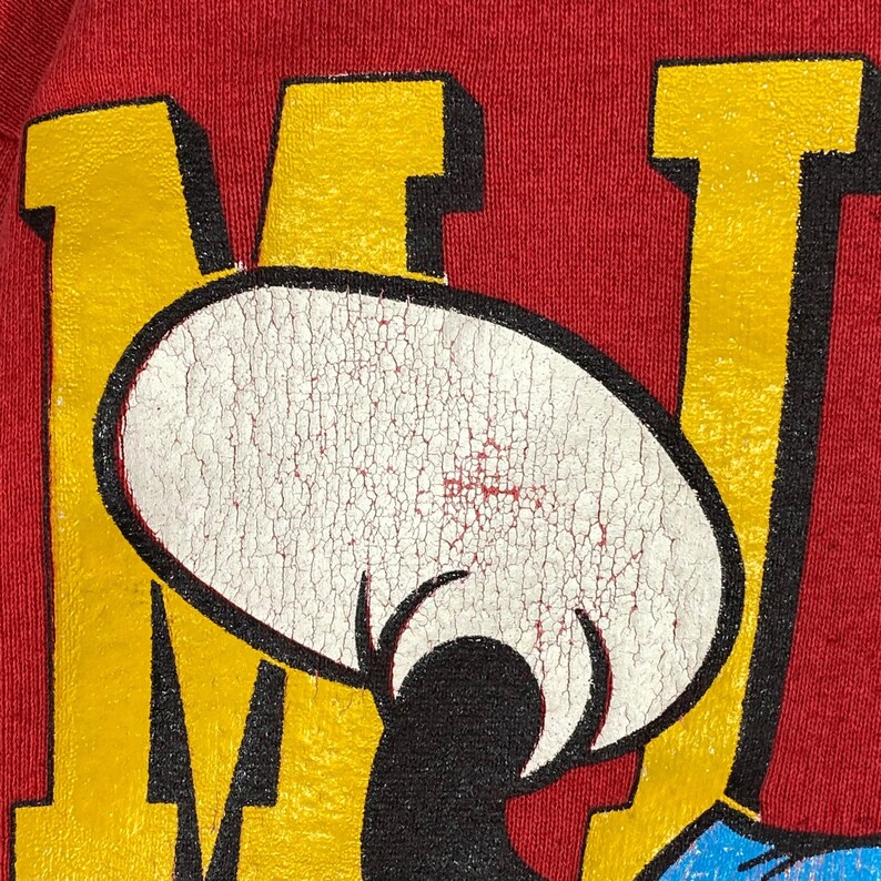 May include: Close-up of a red t-shirt featuring a distressed graphic of a cartoon character's face. The character's face is white with black details, set against yellow and red block letters. The shirt's fabric has a textured appearance.