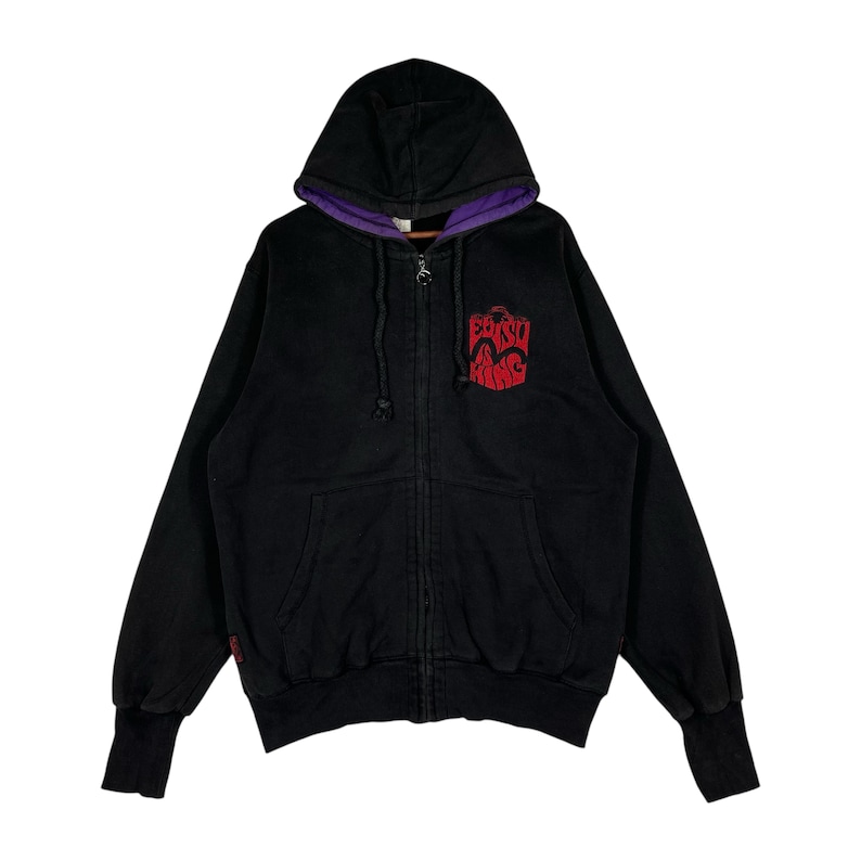 May include: Black zip-up hoodie with a purple-lined hood. The hoodie features a red embroidered logo on the chest. The cuffs and hem are ribbed. The hoodie has a full-length zipper and a drawstring hood.