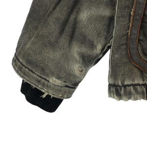 May include: Close-up of a distressed denim jacket sleeve. The jacket is a faded olive green with intentional rips and tears. The cuff is black ribbed knit. Brown leather detailing is visible on the jacket.