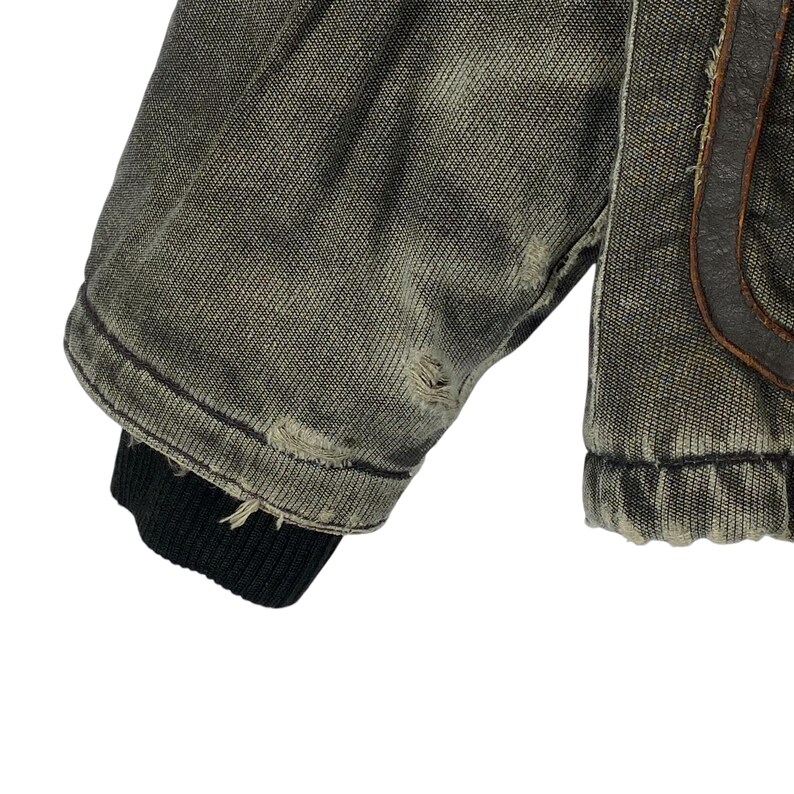 May include: Close-up of a distressed denim jacket sleeve. The jacket is a faded olive green with intentional rips and tears. The cuff is black ribbed knit. Brown leather detailing is visible on the jacket.