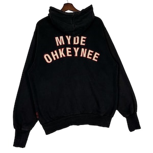 May include: Black hoodie with the text "MYDE OHKEYNEE" in white with red trim across the back. The hoodie has a hood, long sleeves, and ribbed cuffs. The garment is hanging on a wooden hanger.