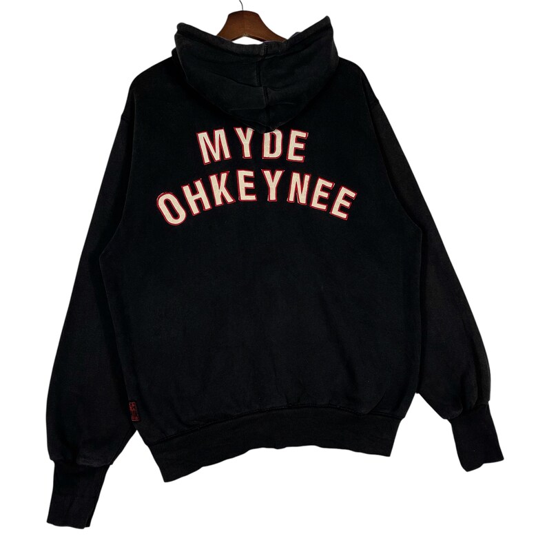 May include: Black hoodie with the text "MYDE OHKEYNEE" in white with red trim across the back. The hoodie has a hood, long sleeves, and ribbed cuffs. The garment is hanging on a wooden hanger.