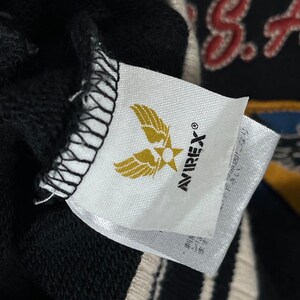 May include: A close-up of a white label with the Avirex logo, a gold emblem of wings and a star. The label is attached to a black garment, with a glimpse of a design in the background.