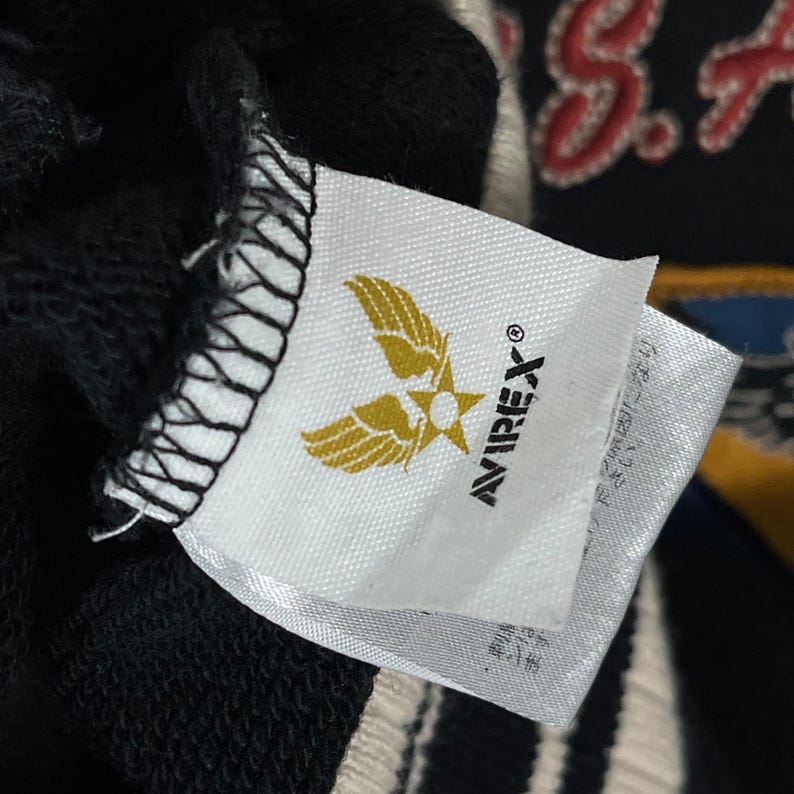 May include: A close-up of a white label with the Avirex logo, a gold emblem of wings and a star. The label is attached to a black garment, with a glimpse of a design in the background.