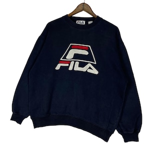 Puede incluir: Sudadera FILA azul marino con logo blanco y rojo. La sudadera tiene cuello redondo y mangas largas. El logo de FILA se muestra prominentemente en la parte delantera. La sudadera est&aacute; colgada en una percha de madera.