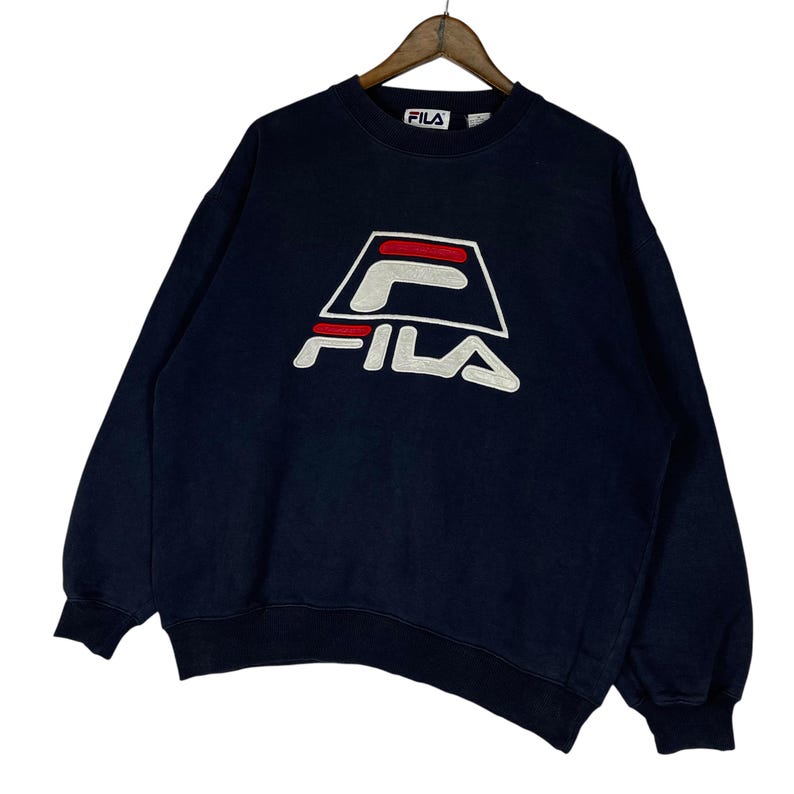 Puede incluir: Sudadera FILA azul marino con logo blanco y rojo. La sudadera tiene cuello redondo y mangas largas. El logo de FILA se muestra prominentemente en la parte delantera. La sudadera est&aacute; colgada en una percha de madera.
