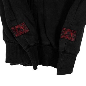 May include: Black sweatshirt with red embroidered patches on the sleeves. The patches feature a stylized face design within a square frame. The cuffs and hem are ribbed. The fabric appears to be a soft, slightly faded material.