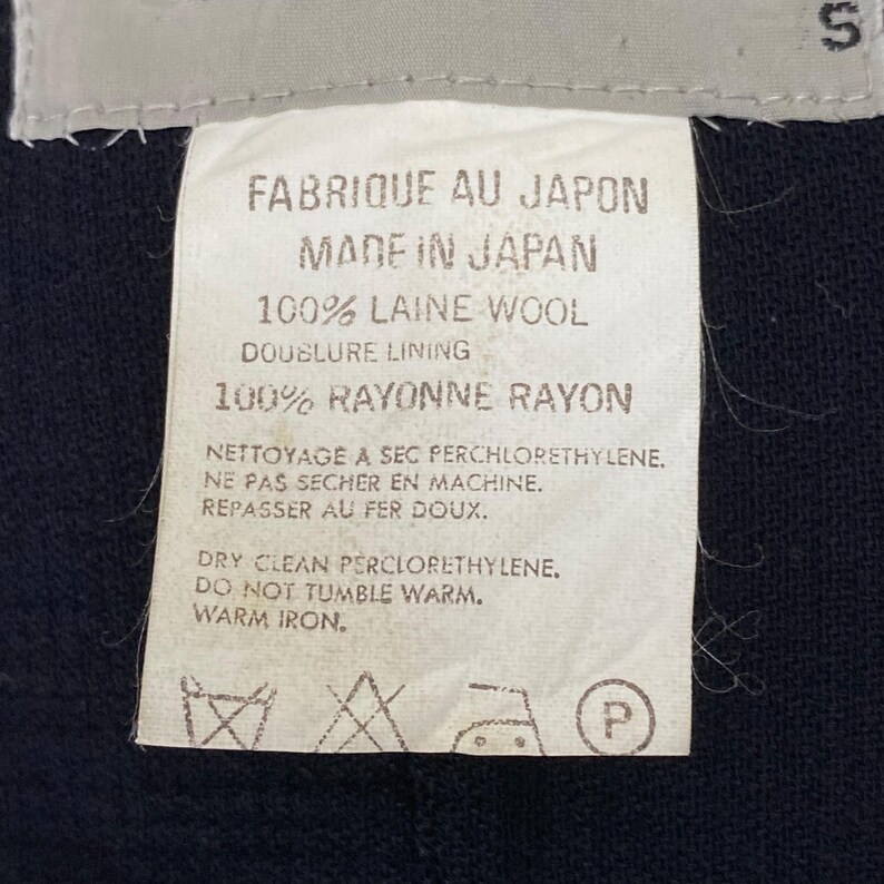 May include: A white fabric tag with black text, including "FABRIQUE AU JAPON" and "MADE IN JAPAN." The tag lists fabric content as 100% laine wool and 100% rayonne rayon, with care instructions for dry cleaning and ironing.