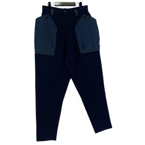 May include: Dark navy blue pants with two large, teal-colored pockets on the front. The pants are hanging on a black hanger. The pockets have a button closure. The pants are made of a solid, dark fabric.