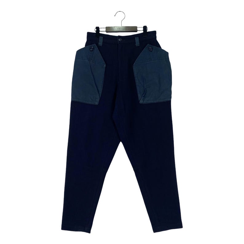 May include: Dark navy blue pants with two large, square, blue-grey pockets on the back. The pants are hanging from a metal hanger. The pockets have a flap and button closure. The pants have a straight leg design.