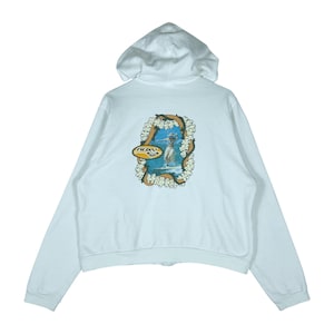 May include: White zip-up hoodie with a hood. The back features a colorful graphic of a surfer with the Roxy logo. The graphic is surrounded by a floral design. The hoodie has long sleeves and a full zipper.