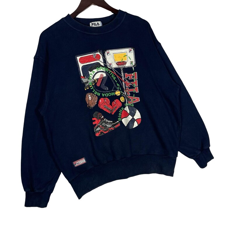 May include: A navy blue crewneck sweatshirt featuring a graphic print of various sports equipment including a basketball hoop, boxing gloves, and a ball. The text "FILA" and "MODA NELLA SPORTIVA" is printed on the front. A small Fila brand tag is on the hem.