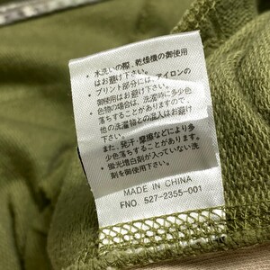 Puede incluir: Primer plano de una etiqueta blanca con texto japon&eacute;s e ingl&eacute;s. La etiqueta est&aacute; adherida a una prenda verde. El texto incluye instrucciones de cuidado y la frase "MADE IN CHINA".