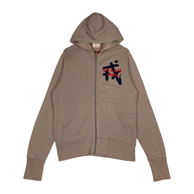 Puede incluir: Una sudadera con capucha con cremallera de color beige con capucha y bolsillos delanteros. La sudadera con capucha presenta el texto "E.V.I.S.U. TRACK" en rojo y azul marino en la parte delantera. Los pu&ntilde;os y el dobladillo son acanalados.