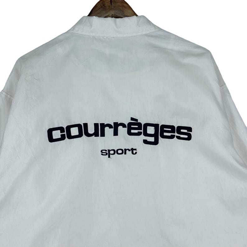 May include: White jacket with the black text "courr&egrave;ges sport" on the back. The jacket is hanging on a wooden hanger. The jacket has a collar and a button-up front.
