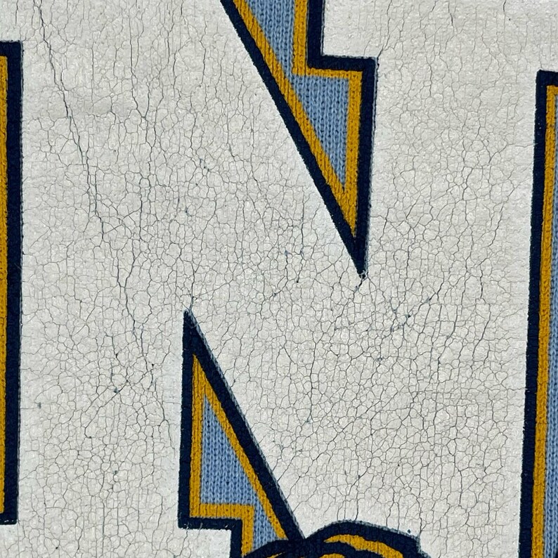 May include: A close-up view of a white graphic print on a light blue fabric background. The print features a large letter N with a dark blue outline and a yellow inner border. The white ink shows significant cracking and a weathered texture.