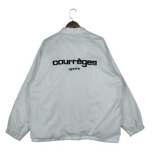 May include: White jacket with the text "courr&egrave;ges sport" in black on the back. The jacket has a collar and long sleeves. The jacket is hanging on a wooden hanger.